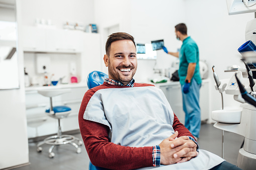 Male dental patient, sitting in a dental chair smiling with a technician looking at a computer behind him